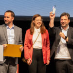 Drei Personen, ein Mann hält Preis in die Höhe