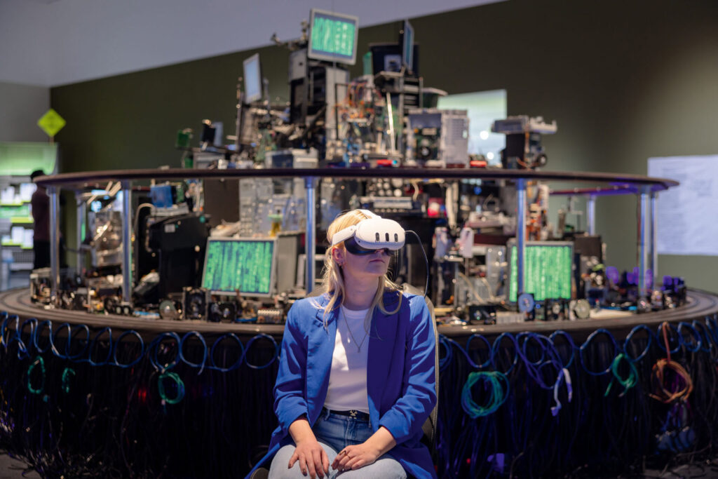Frau mit VR-Brille vor einer Installation aus elektronischen Geräten
